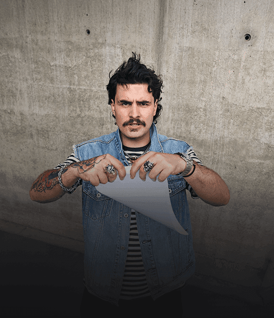 Mężczyzna z wąsem i tatuażami rozdziera kartkę papieru, patrząc poważnie w stronę obiektywu