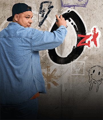 Mężczyzna w czapce z daszkiem i jeansowej kurtce maluje graffiti na betonowej ścianie