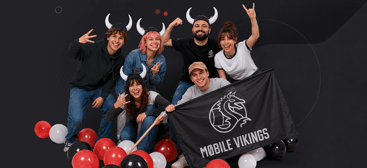 Grupa radosnych osób w hełmach z rogami pozuje wśród balonów z flagą Mobile Vikings